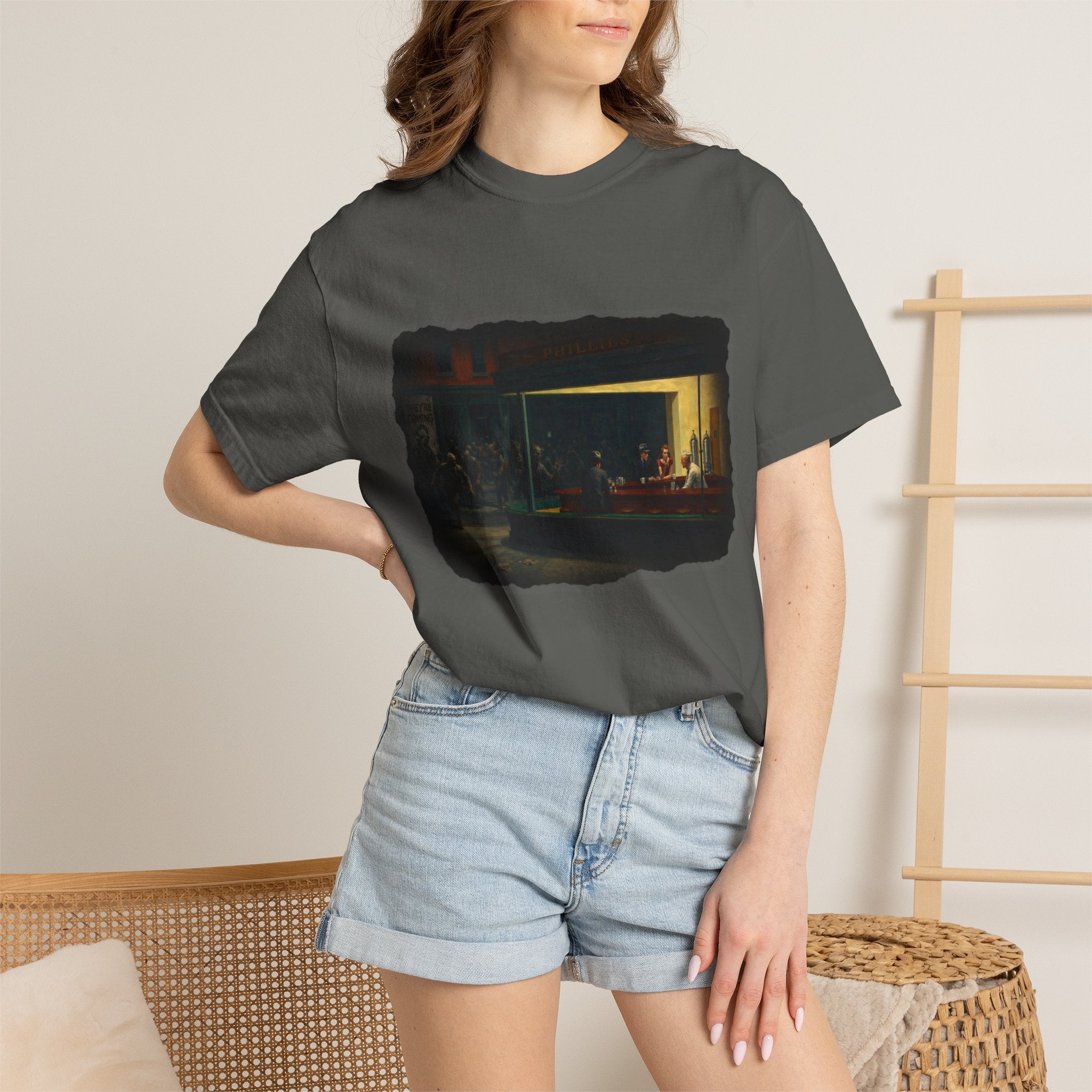Woman standing by a wicker chair wearing pepper colour tshirt with graphic image inspired by Edward Hopper's Nighthawks 