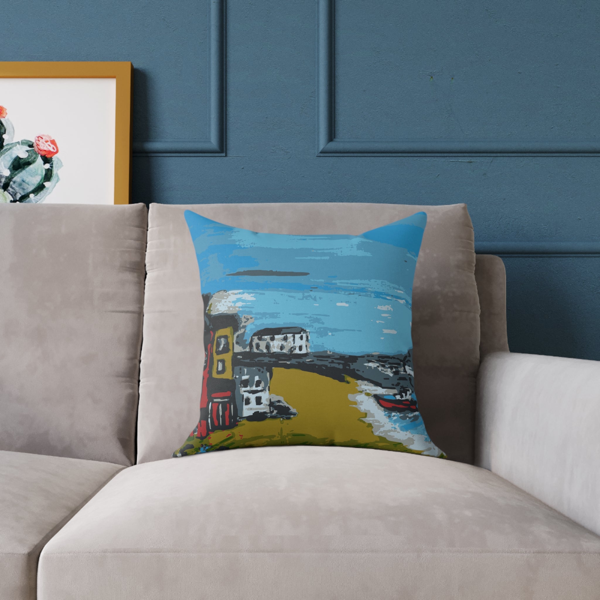 Decorative pillow with a colorful coastal scene on a beige sofa against a blue wall.