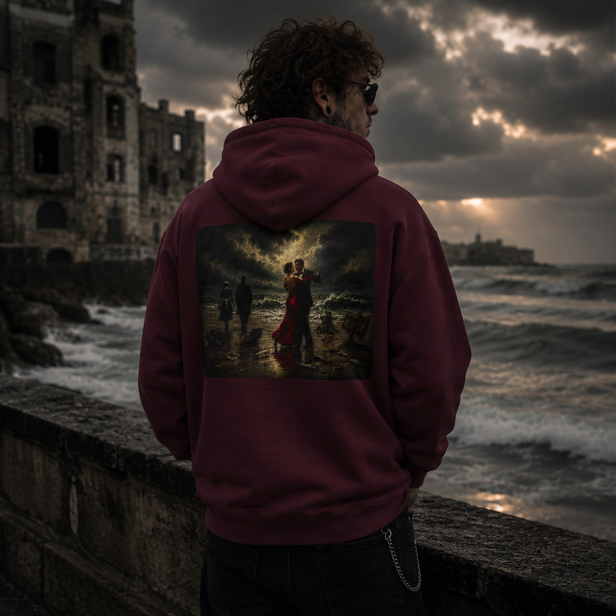 Man in sunglasses standing against a sea wall in stormy weather wearing a maroon hoodie with a graphic of a Vettriano inspired Singing Butler print on the back 